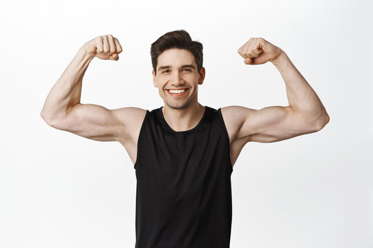 Sport and gym. Smiling young healthy man flexing biceps, showing strong big muscles and fit body, standing in tank top over white background