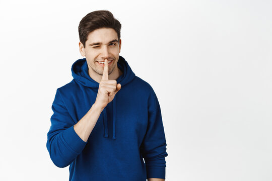 Handsome Smiling Man Winks And Shows Shh, Shush Silence Gesture, Press Finger To Lips, Hiding Secret, Prepare Surprise, Stands Over White Background