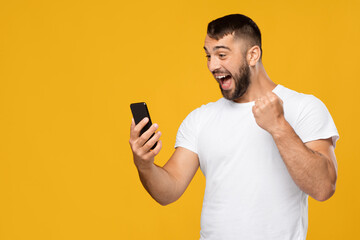 Satisfied excited adult european guy in white t-shirt looking at smartphone and expresses joy, celebrating success