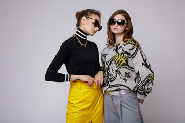 Two fashion models in gray pants, black blouse, yellow leather long skirt, stylish sunglasses. Beautiful young women. Studio shot, portrait. White gray background.