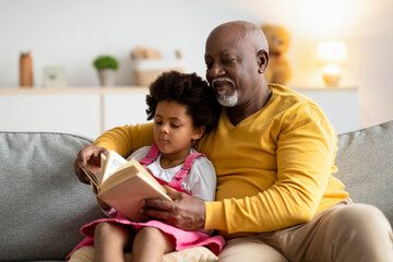 Concentrated african american small curly girl and smiling elderly man reading book with fairy tales