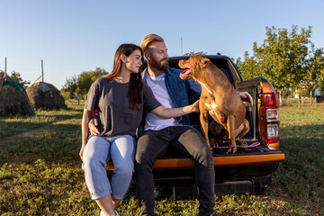 Young couple sitting on the back of a pickup truck with their trustful guardian a canine American bully - obrazy, fototapety, plakaty
