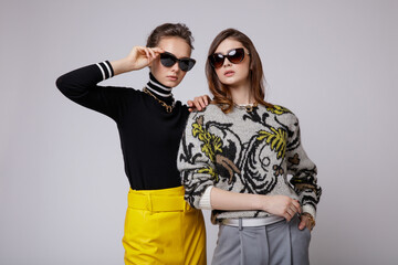 Two fashion models in gray pants, black blouse, yellow leather long skirt, stylish sunglasses. Beautiful young women. Studio shot, portrait. White gray background.