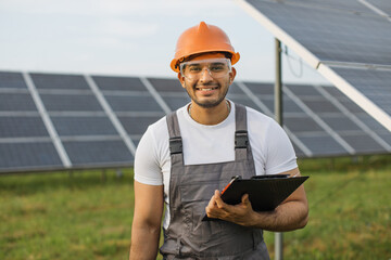 Handsome smiling man in uniform, orange hard hat and safety glasses holding clipboard and instruments for work on solar farm. Competent energy engineer controlling work of photovoltaic cells.
