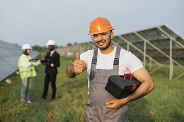 Arabian technician in orange helmet showing thumb up and smiling on camera while two engineers talking behind. Multiracial people working on big station with solar panels.