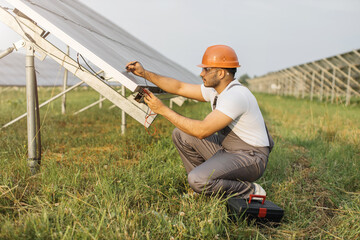 Indian man in uniform squatting near solar panel with multimeter in hands and smiling on camera....