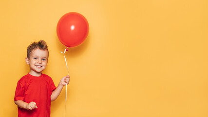A happy Caucasian boy isolated on a bright yellow background holds a red balloon in his hands. A place for your text or advertisement. - Powered by Adobe