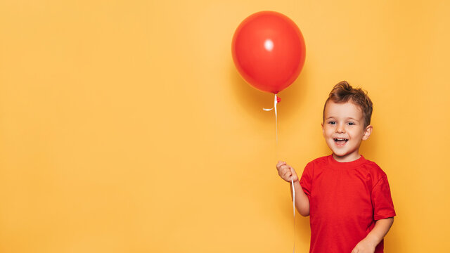 A happy Caucasian boy isolated on a bright yellow background holds a red balloon in his hands. A place for your text or advertisement.