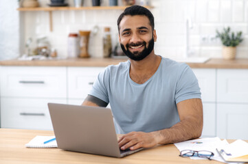 Obraz na płótnie Canvas Portrait of a positive friendly bearded Indian man, sitting at home at table in kitchen, working in laptop remotely from home, looking directly at camera and smiling