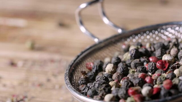 Macro shot of sieve with mix of peppercorns falling on table in restaurant kitchen. Pink, white and black fragrant peppers peas close up. Falling colorful seeds of hot peppercorns in slow motion.