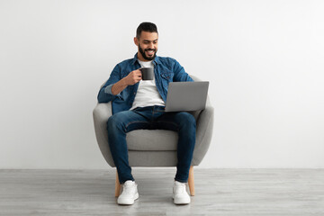 Cheerful young Arab man working online, sitting in armchair with coffee and using laptop against white studio wall