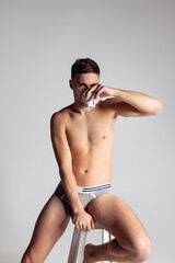 Portrait of young handsome shirtless man sitting on high chair isolated on gray studio background. Natural beauty of male body