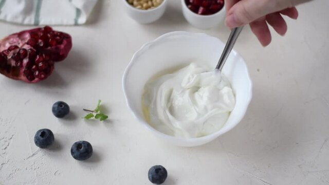 Mixing Greek yogurt in the white ceramic bowl with spoon before adding berries and nuts. Thick dairy product and toppings around. Making healthy breakfast