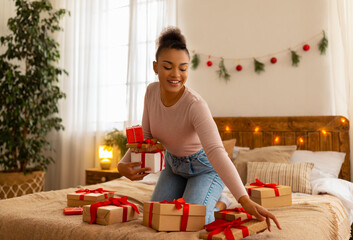 Positive african american woman holding presents in hands, sitting on bed surrounded by Christmas gifts and smiling