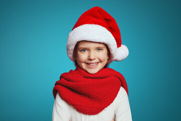 Cheerful little girl wearing christmas hat and red scarf, looking at camera while standing isolated over blue background 