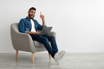 Millennial Arab man having creative idea, gesturing eureka, sitting in armchair with laptop against white studio wall