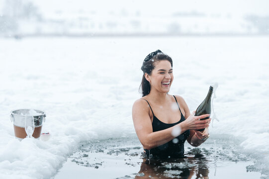 Girl with champagne in frozen lake ice hole. Woman hardening the body in cold water. Successful woman concept