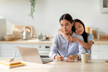 Family business. Young asian woman working on laptop and taking notes, cute daughter embracing mother at home