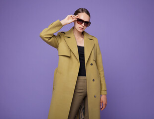 Fashion photo of a beautiful elegant young woman in a pretty yellow mustard coat, pants, trousers posing on a purple lavender background. Studio shot, portrait. Stylish sunglasses