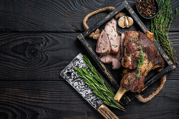 Roasted mutton lamb leg sliced in a wooden tray with meat cleaver. Black wooden background. Top...
