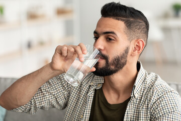 Fototapeta na wymiar Healthy lifestyle concept. Millennial Arab guy drinking water from glass at home