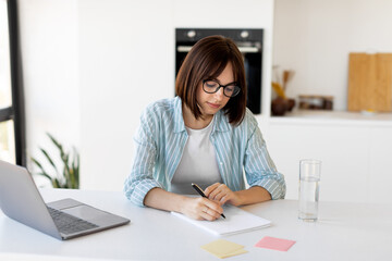 Millennial female student studying with laptop and taking notes, sitting in kitchen, watching webinar on computer