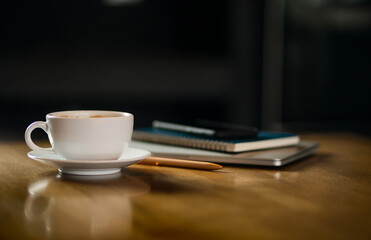 Closeup white cup of coffee with smartphone, notebook and pen on wooden table in cafe