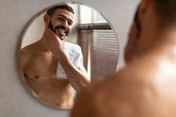Confident young Arab guy looking in the mirror, touching chin