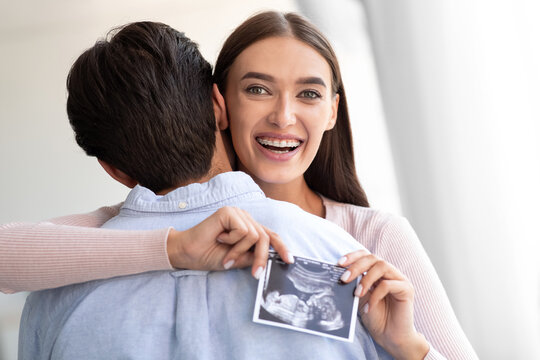 Happy european young wife hugs husband and holds ultrasound picture of baby in living room interior