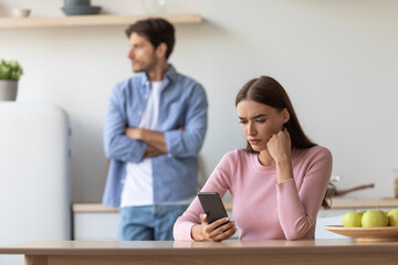 Unhappy offended young caucasian husband ignoring wife, lady chatting on smartphone