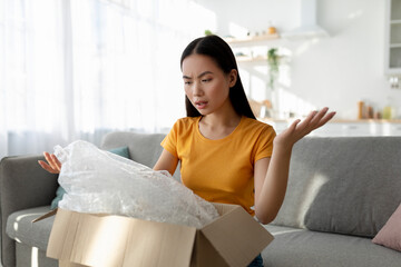 Terrible delivery service. Unhappy asian woman unboxing cardboard parcel, receiving damaged item,...