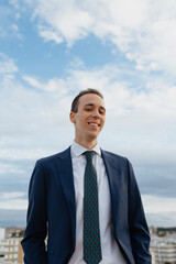 Young businessman in a suit and tie looking at camera and smiling while standing outside in a rooftop with a cloudy sky