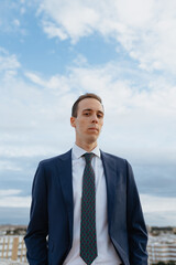 Young businessman in a suit and tie looking serious at camera standing outside in a rooftop with a cloudy sky