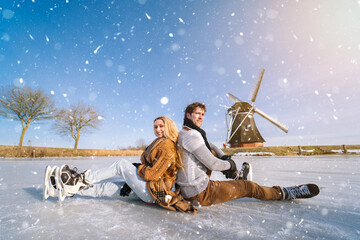 Fototapeta na wymiar Loving couple having fun on ice in typical dutch landscape with windmill. Woman and man ice skating outdoors in sunny snowy day. Romantic Active date on frozen canal in winter Christmas Eve.