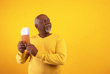 Happy senior black man holding passport with plane boarding tickets, looking aside at empty space on orange background