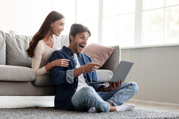 Happy surprised excited european young couple show fingers to laptop rejoice to victory in living room