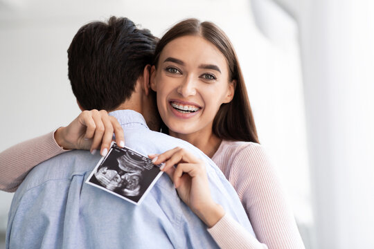 Cheerful millennial caucasian wife hugging husband and holding ultrasound picture of baby in living room