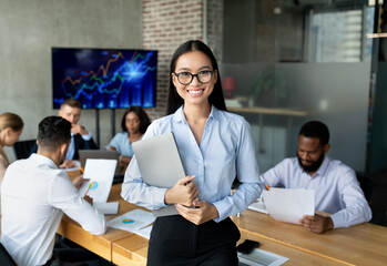 Smiling Asian Female Employee Posing In Office Interior During Meeting With Colleagues