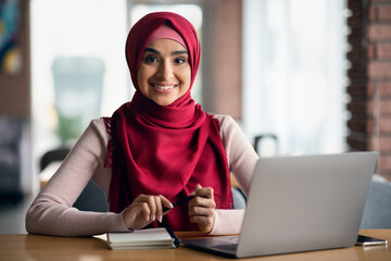 Cheerful muslim woman sitting at table in front of laptop