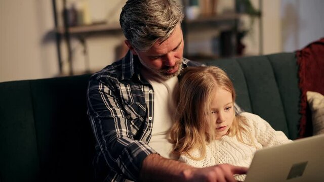 Attractive father checking homework, helping cute daughter with studies sit at sofa. Father and kid e learning together having remote distant lesson. High quality 4k footage