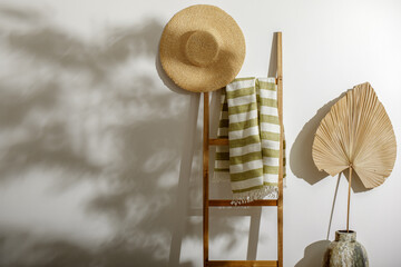 Organic linen beach towel on wood ladder, straw hat and dried palm leaf against white wall with...