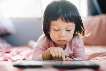 A little girl in a pink shirt lay intently playing the tablet on her bed. self-learning technology education concept