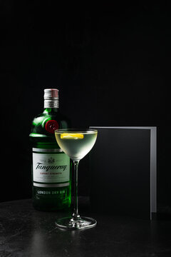 Minsk/Belarus - 11-24-2021: An alcoholic cocktail in a nick and nora glass garnished with fruit slice, standing on an opened menu book on black wooden table, and a bottle of Tanqueray gin
