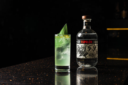 Minsk/Belarus - 11-24-2021: A light green alcoholic cocktail in a highball glass with ice, garnished with a bamboo leaf, at the bar counter, and a bottle of Tequila Espolon Blanco