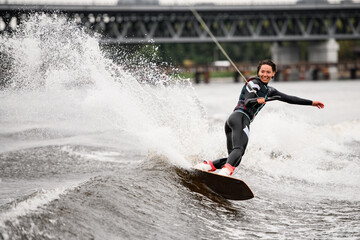 cheerful young woman rides down splashing wave on wakeboarding board holding rope with her hand