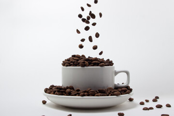Coffee beans fall into the cup. White cup overflowing with coffee beans on a white background....