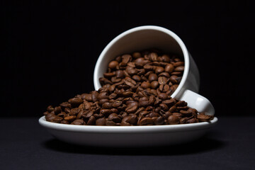 Cup filled with coffee beans on a black background. Freshly roasted coffee beans are poured out of the cup. Excessive consumption of caffeine