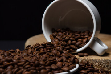 Cup filled with coffee beans on a black background. Freshly roasted coffee beans are poured out of the cup. Excessive consumption of caffeine