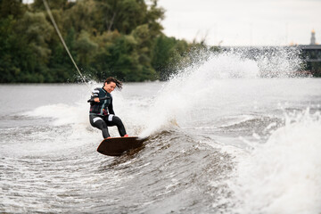 active female wakeboarder balances on splashing wave on board holding rope with her hand