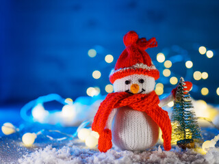 Christmas card with a knitted snowman at the Christmas tree. Fake snow, blue background